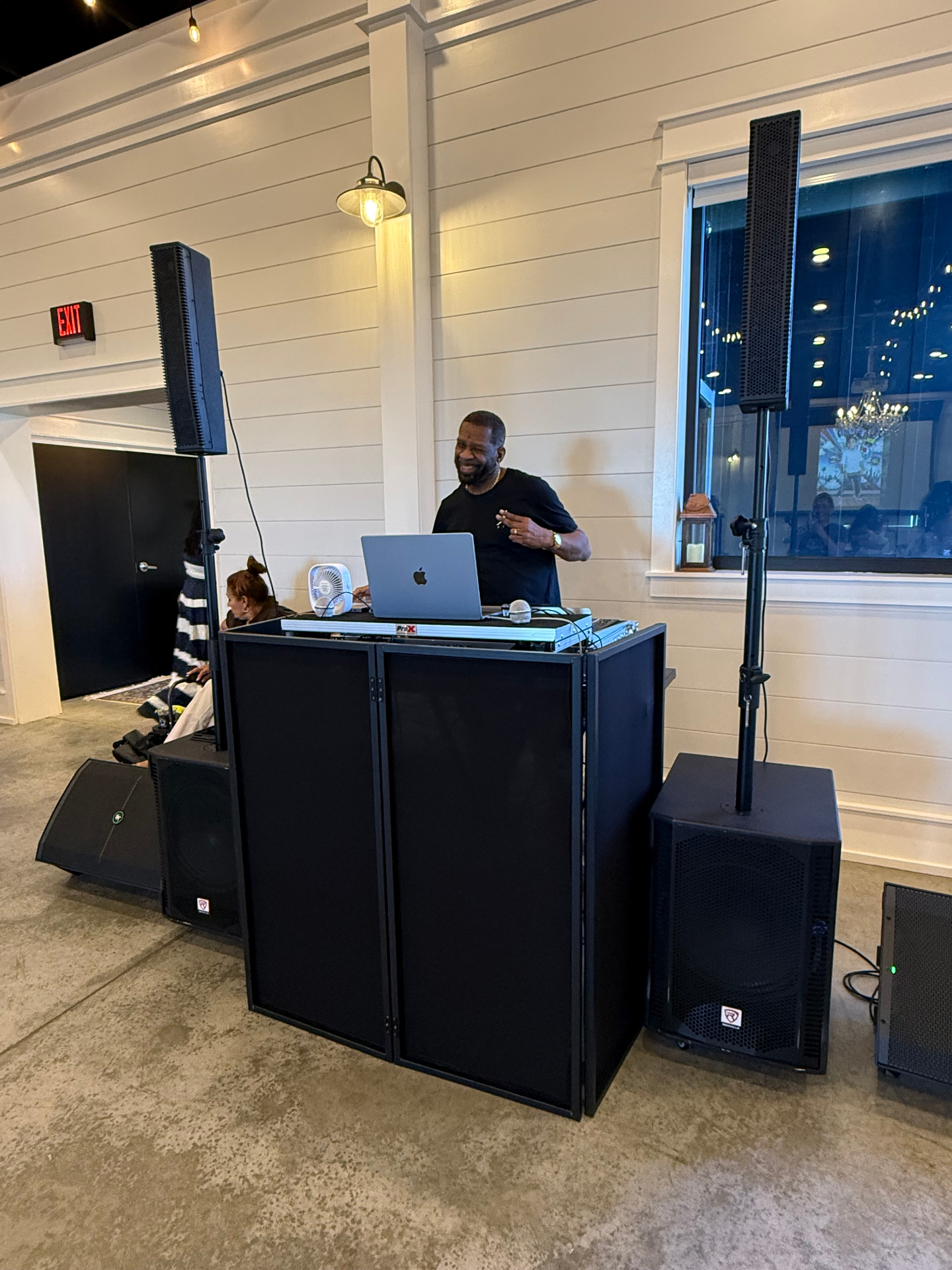 Independent DJ setup with column line array speakers, subwoofers, and a black facade booth at an indoor event venue