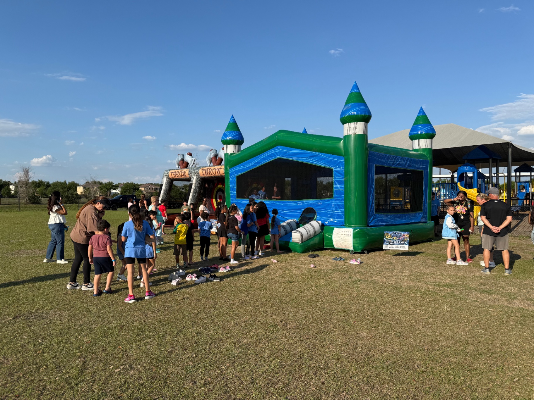 Crowd of elementary students lined up barefoot to enter a green and blue castle bounce house at a school field day, with a throw-game inflatable in the background