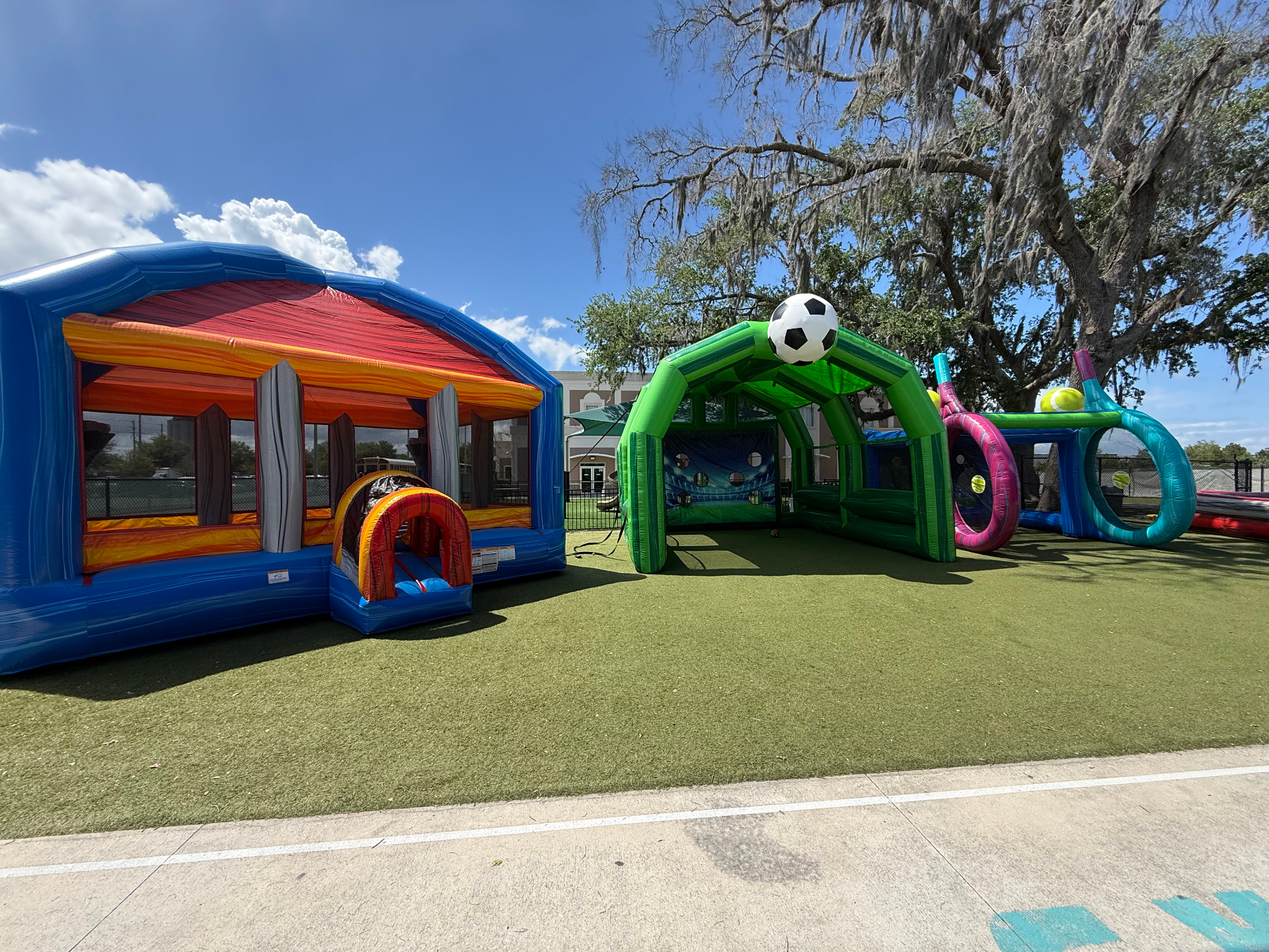 School field day setup with a sports arena bounce house, inflatable soccer kick game, and giant tennis ring toss on turf