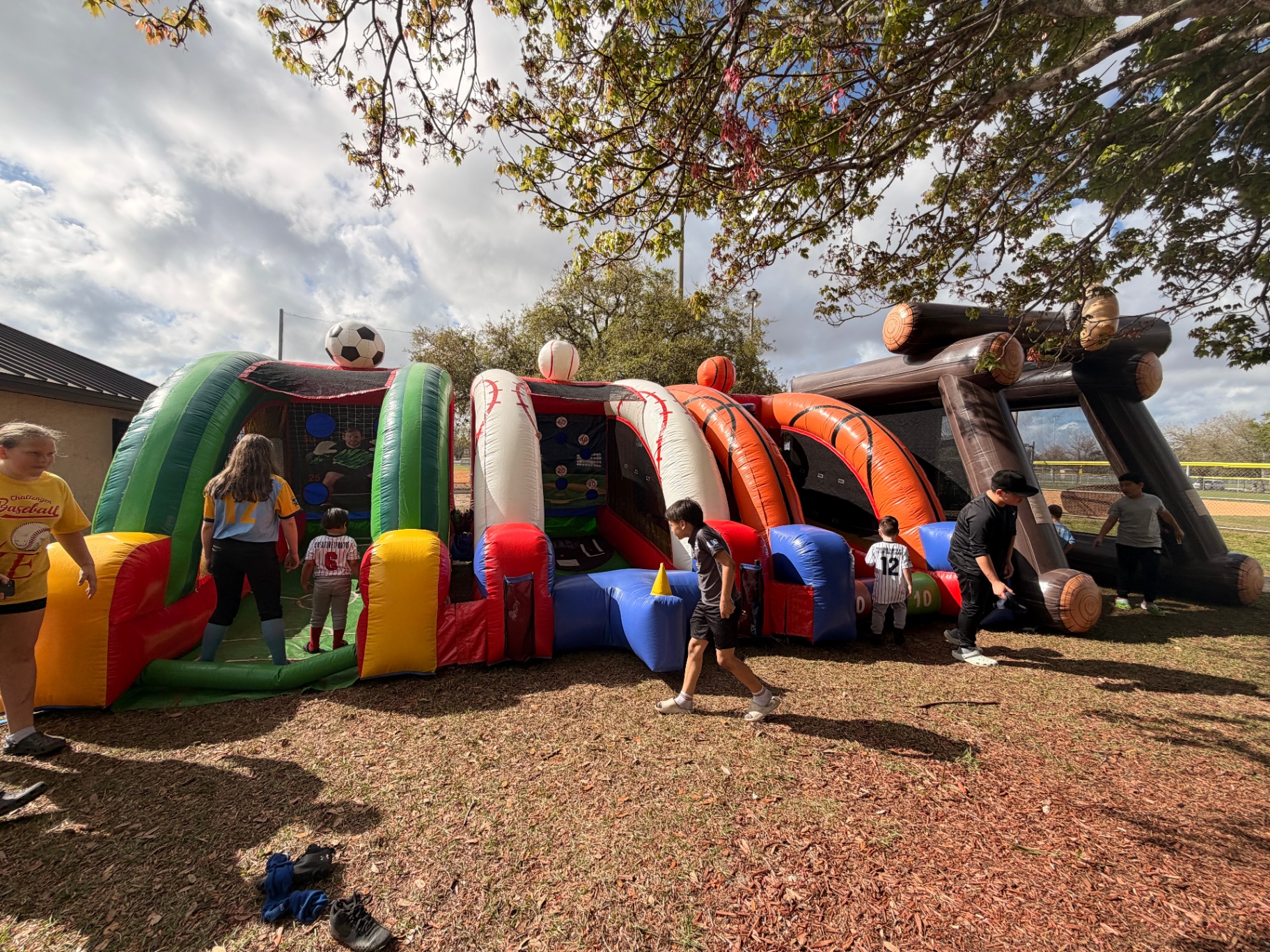 Multi-sport inflatable interactive game with soccer, baseball, and basketball lanes set up at a Little League event under a shade tree