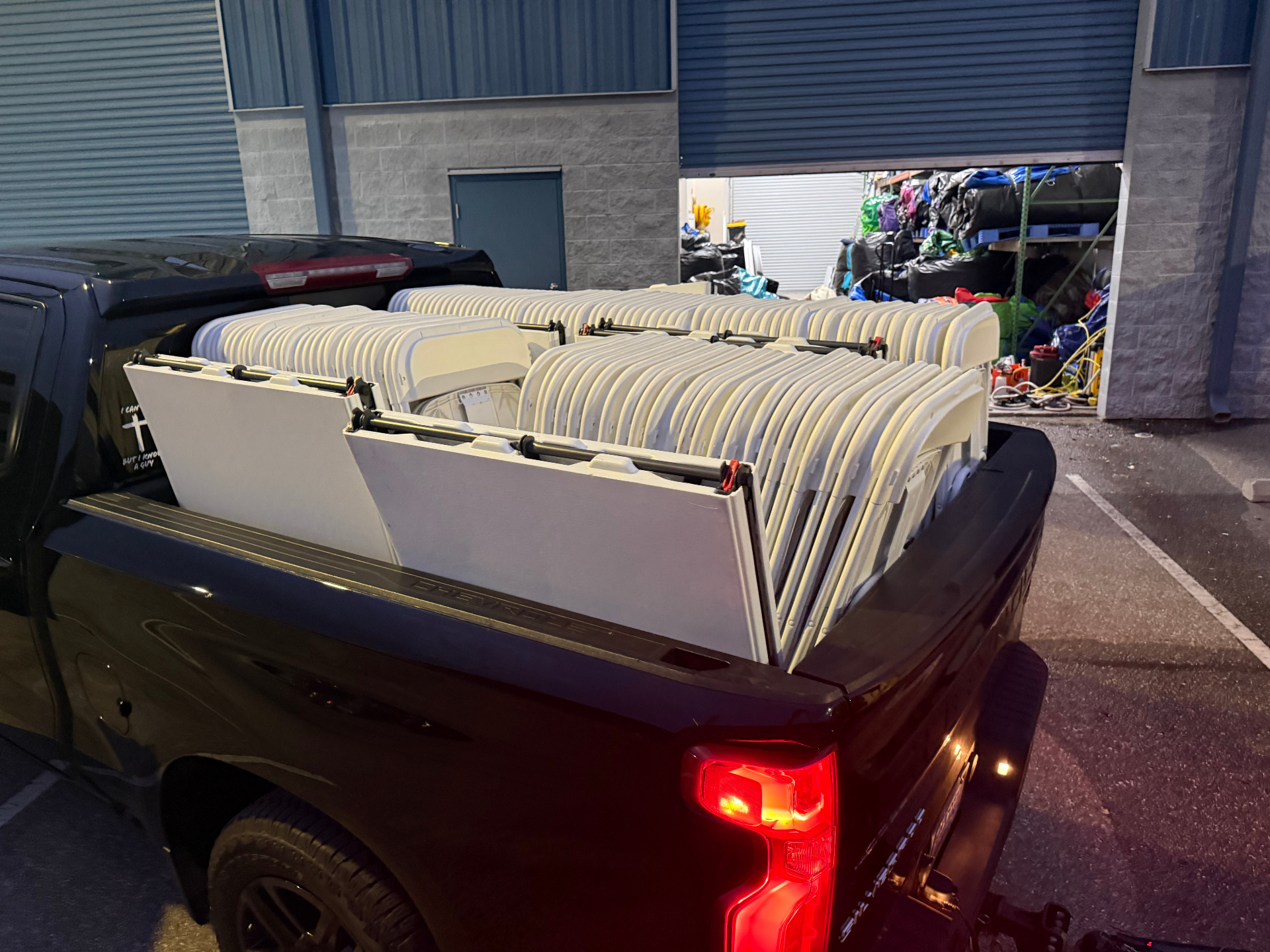 Stack of white folding chairs loaded sideways in the bed of a pickup truck outside a rental warehouse at dusk
