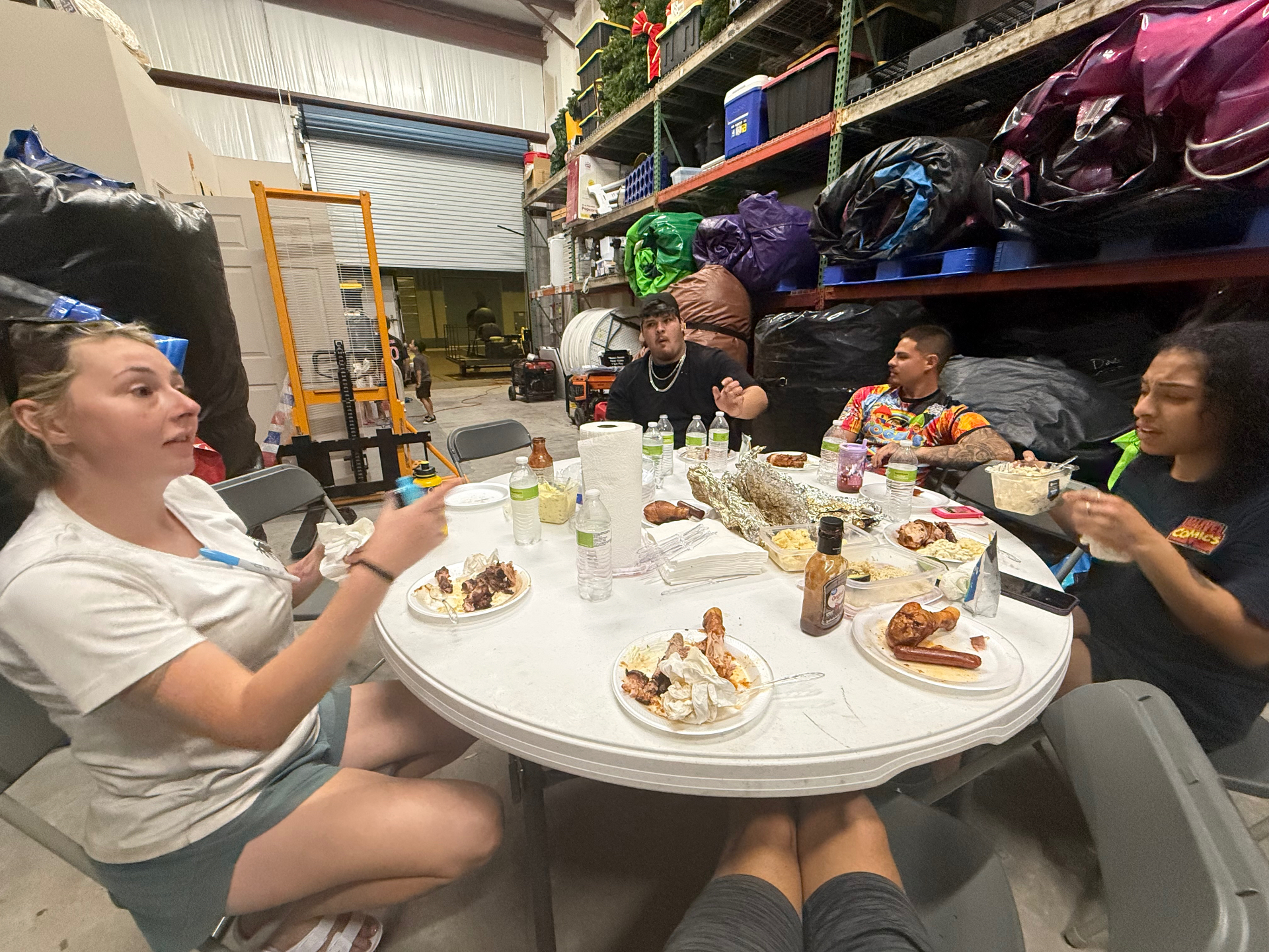 Party rental crew gathered around a folding table inside the warehouse, sharing a barbecue dinner together after a long day of events.