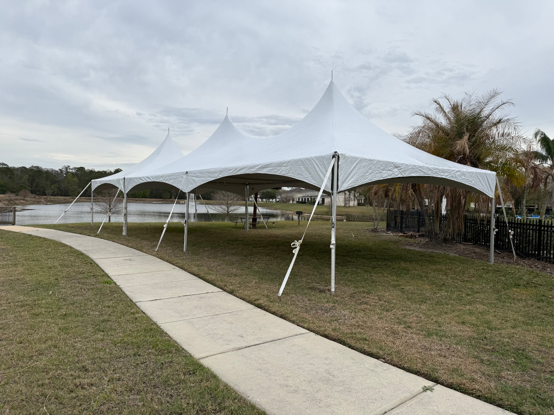 Three white high-peak frame tents installed end-to-end on a lakeside lawn for a wedding event, with palm trees and a clubhouse in the background